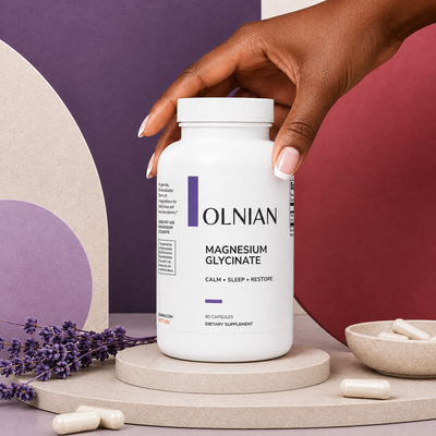 A white bottle of OLNIAN Magnesium Glycinate dietary supplement, styled on a round pedestal with lavender sprigs, loose capsules, and a small bowl of capsules against a geometric purple and dusty rose background.