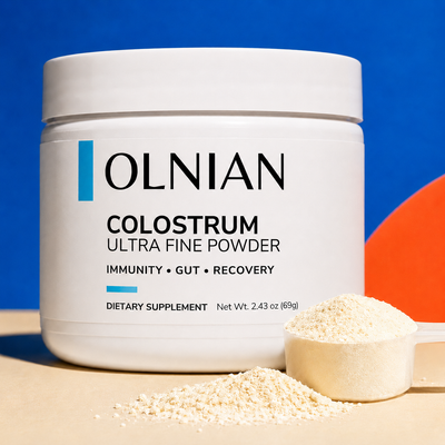 A white plastic tub of OLNIAN Colostrum Powder dietary supplement, shown with a scoop and loose powder in front, set against a blue background with a beige surface.